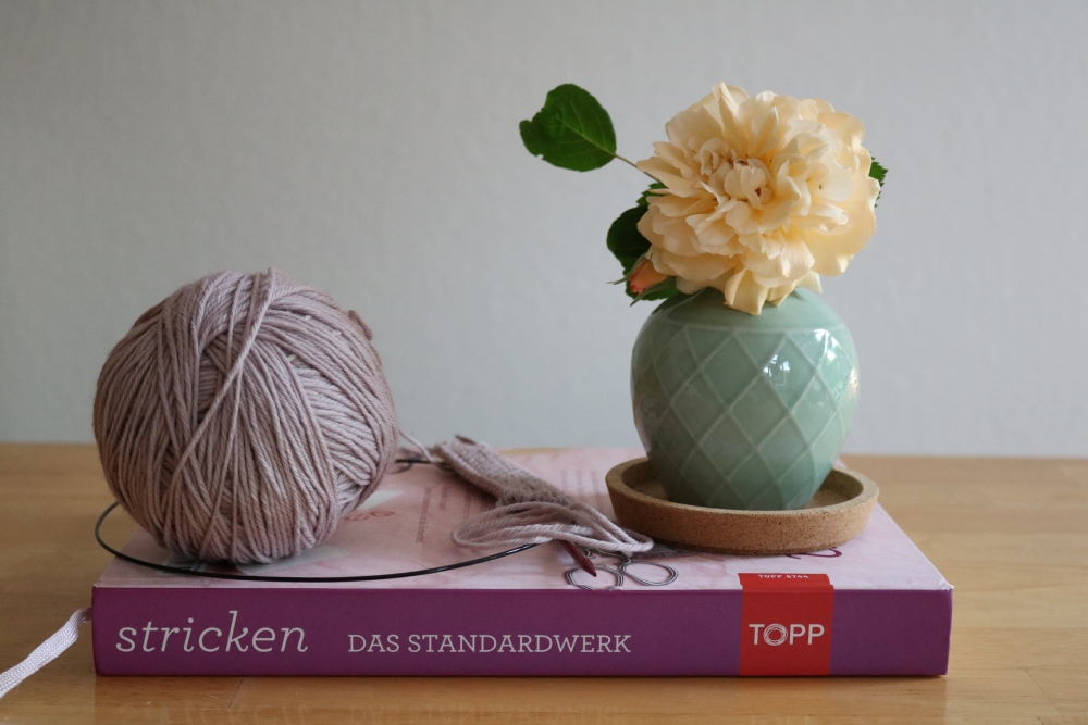 A ball of yarn with circular knitting needles and a small vase with a light yellow flower placed on top of a book of knitting basics
