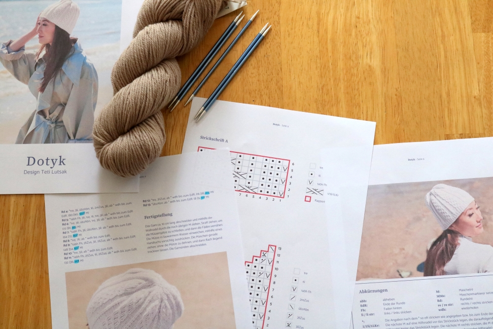 Different pages of knitting pattern for the hat Dotyk with markings and a skein of yarn on a wooden table
