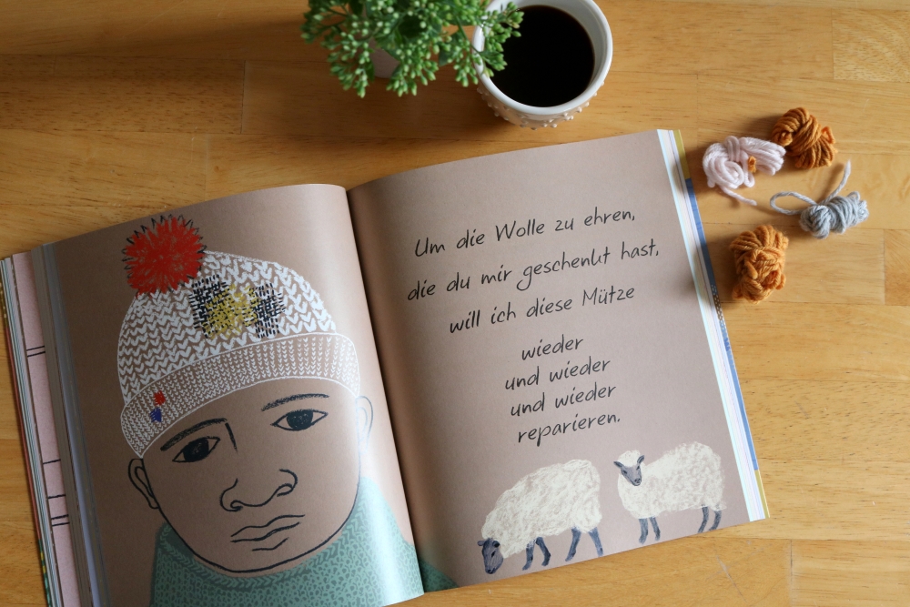 Left-over yarns and an open book on a table. On the right page with sheep drawings you can read in German: To honour the wool you gave me, I will repair this hat again and again and again.