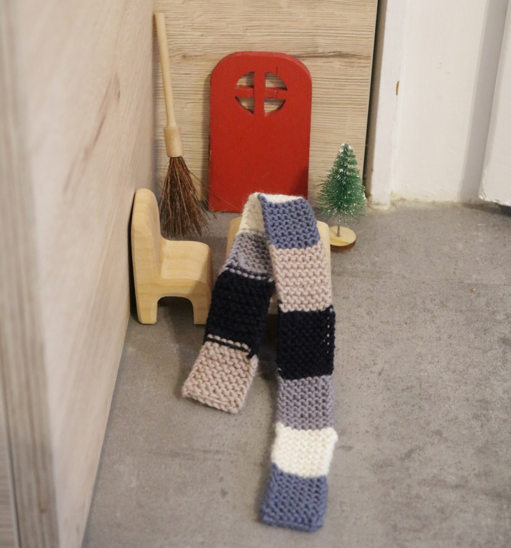 A doll-house table and chair in front of a little red door on the wall, on the table lies a tiny knitted scarf with broad stripes in blues, greys and beige