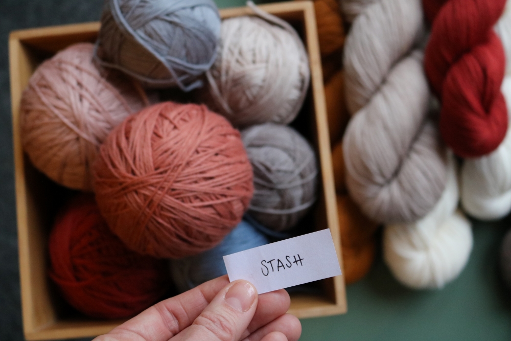Topview: Balls of yarn in a box, next to them some skeins, all slightly blurred. Focused, a hand holding a small piece of paper with the word stash handwritten on it