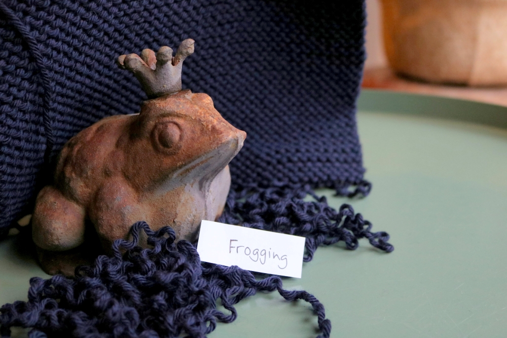 A knitted piece in dark blue, some unravelled yarn coming from it; in between the yarn a terracotta frog and a small piece of paper with the word frogging handwritten on it